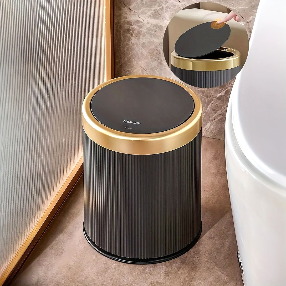 Black Elegance Oval Waste Solution trash can with a press-top lid and gold rim, positioned next to a toilet in a modern bathroom setting. An inset shows a close-up of the lid opening.