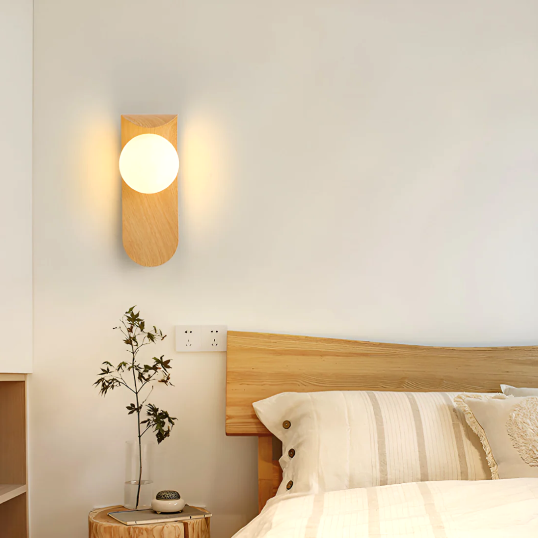 A Nordic Glow LED wall sconce in light wood, emitting a soft light, mounted above a bed in a minimalist bedroom setting with a plant on a wooden stump nightstand.