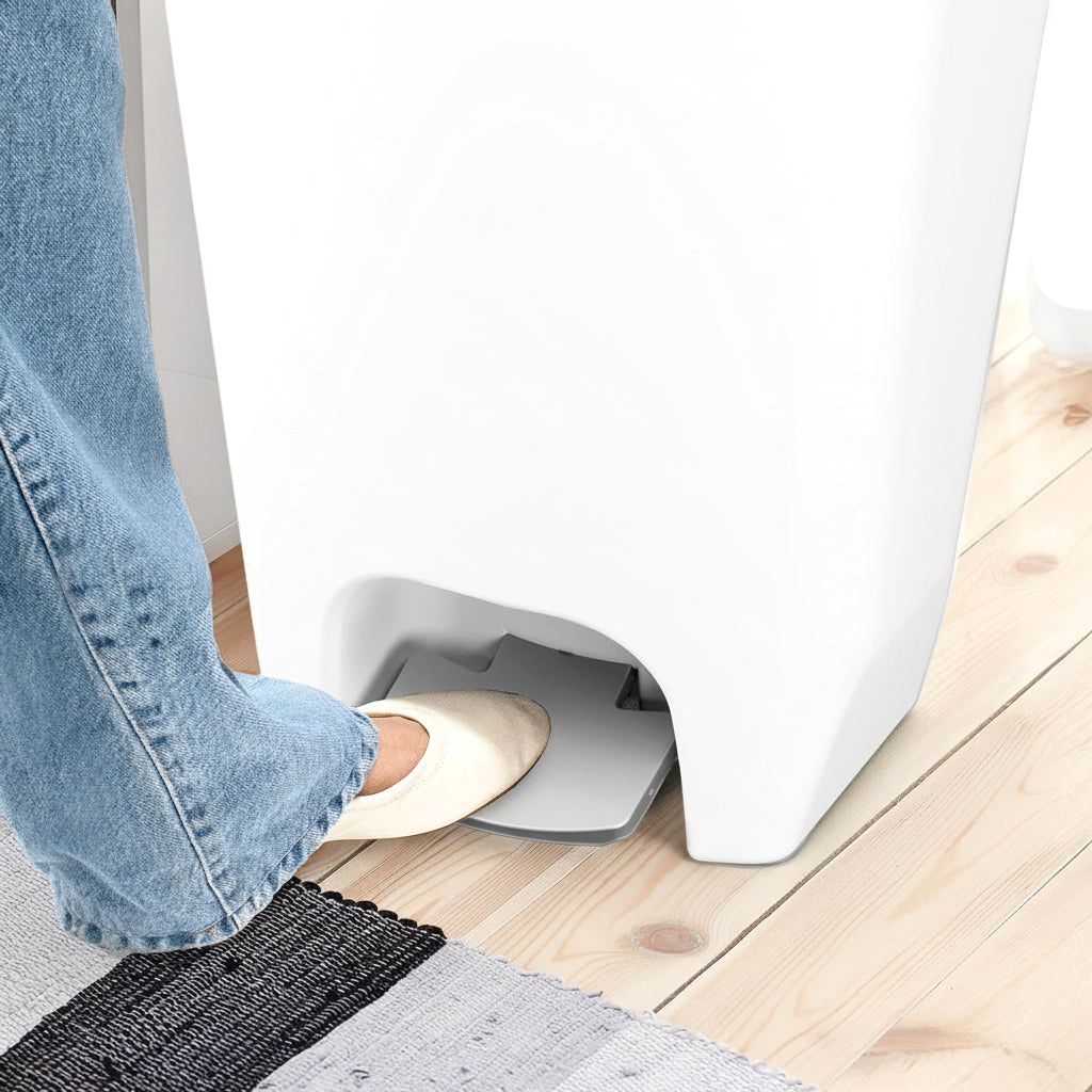 A foot in a light flat shoe pressing the step pedal of a white Serenity kitchen trash can, showcasing hands-free use on a wooden floor with a rug.