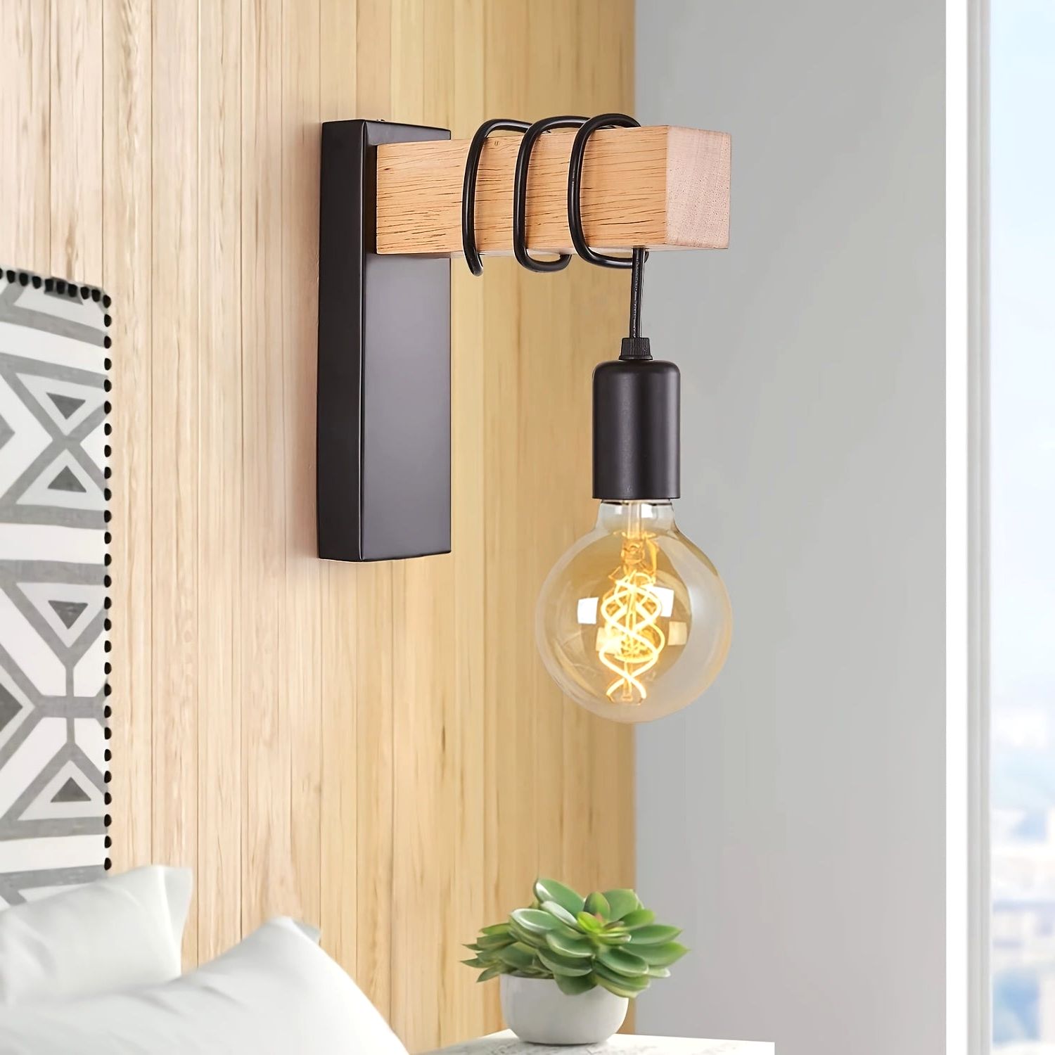 The Urban Forge wall sconce in black metal finish with a wooden block and black coiled accents, holding an exposed E27 bulb with a spiral filament. The sconce is mounted on a wooden paneled wall next to a bed with white pillows and a small plant, highlighting its modern industrial aesthetic.