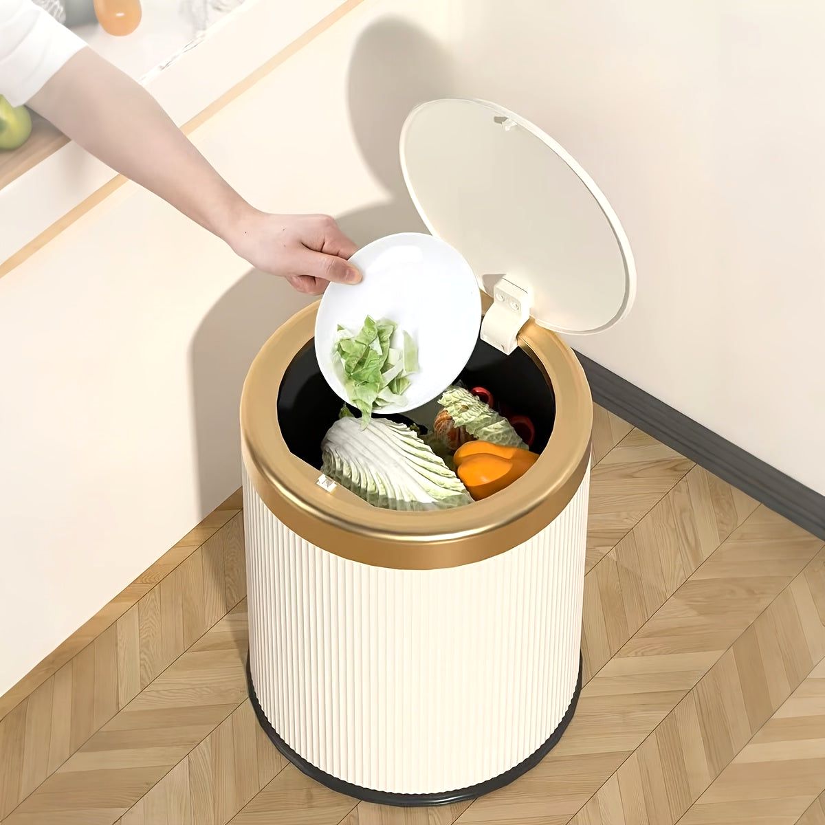 Cream Elegance Oval Waste Solution trash can with a press-top lid and gold rim, shown in a kitchen setting with a hand dropping food scraps from a plate into the open bin.