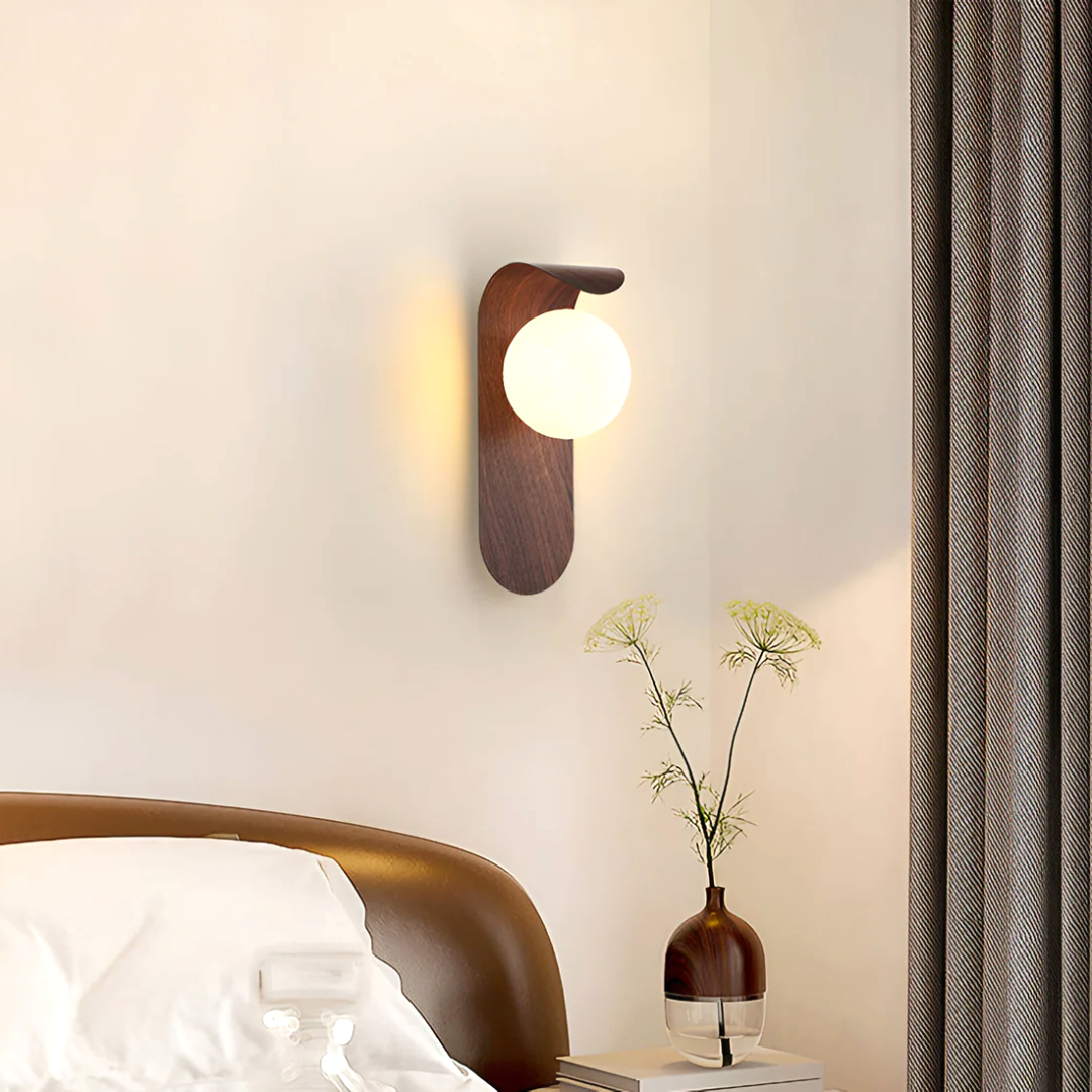 A Nordic Glow LED wall sconce in walnut wood, emitting a soft light, mounted above a bed in a minimalist bedroom setting with a plant on a nightstand.