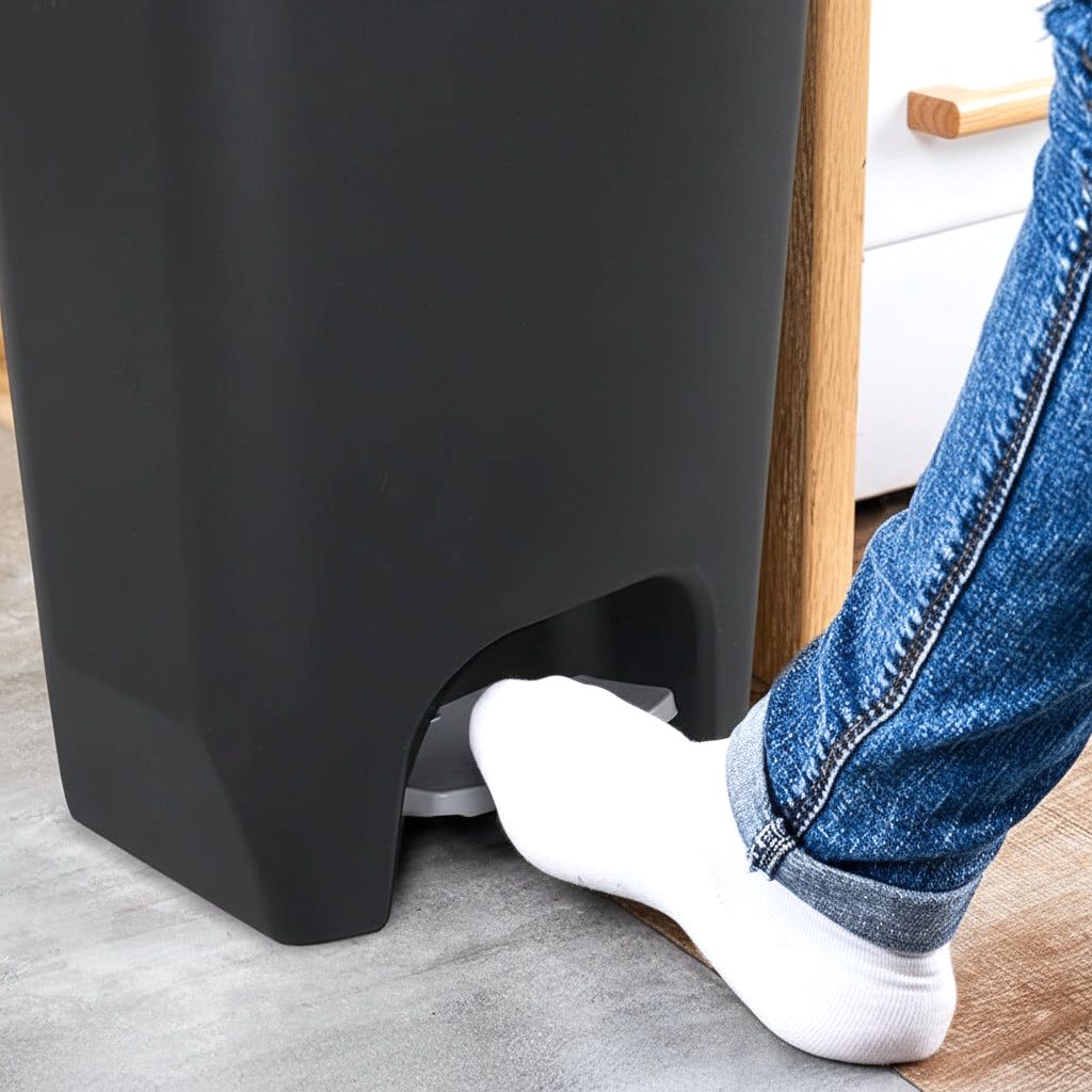 Close-up of a foot in a white sock pressing the step pedal of a black Serenity kitchen trash can, demonstrating hands-free operation.