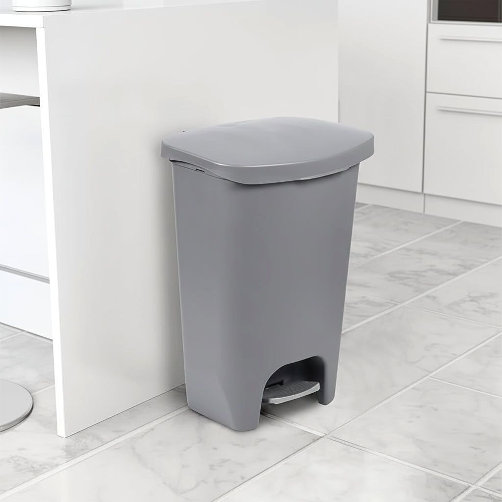 A sleek grey Serenity kitchen trash can with a step pedal, positioned neatly beside a white kitchen island on a light tiled floor.